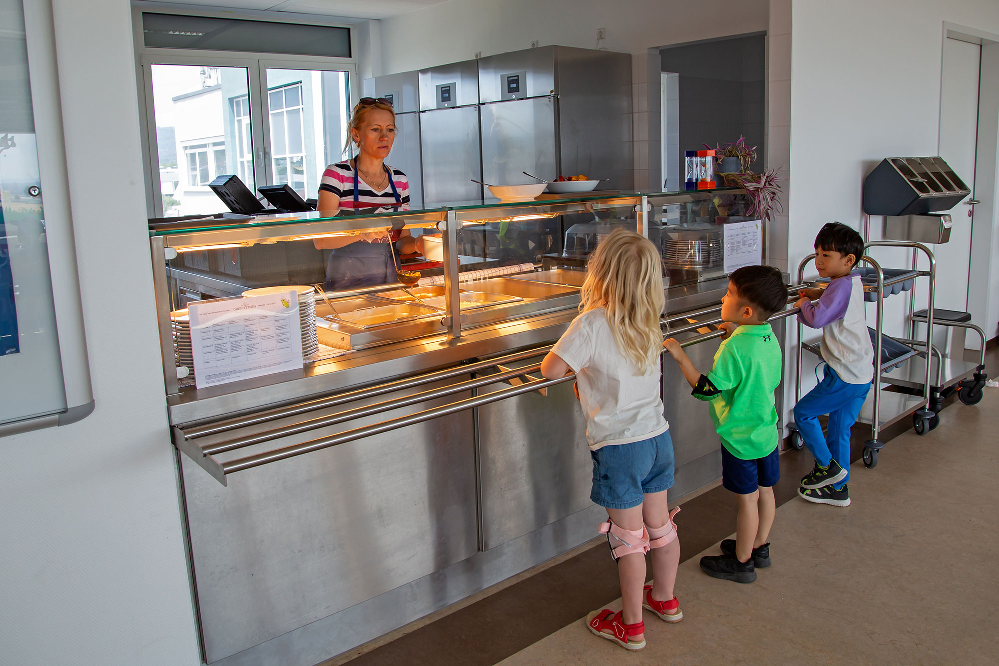 Three kids are standing in a line for a meal in the canteen.