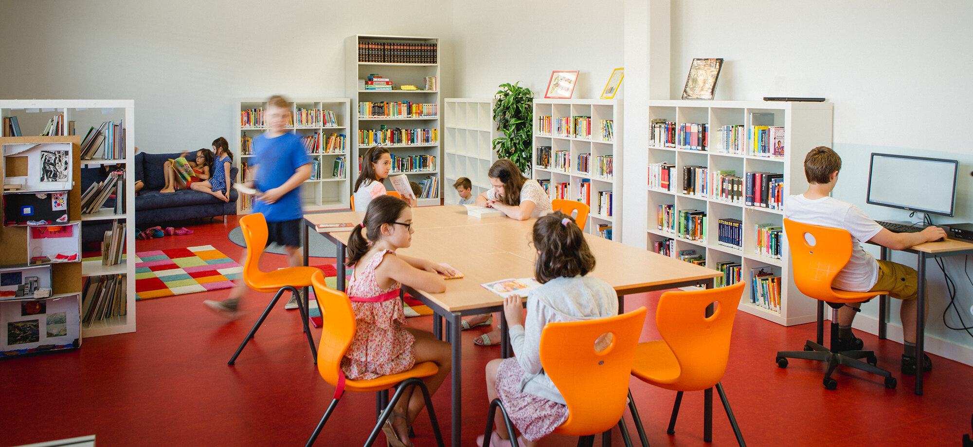 View of the school library. Children are reading, picking up books or sitting at the computer.