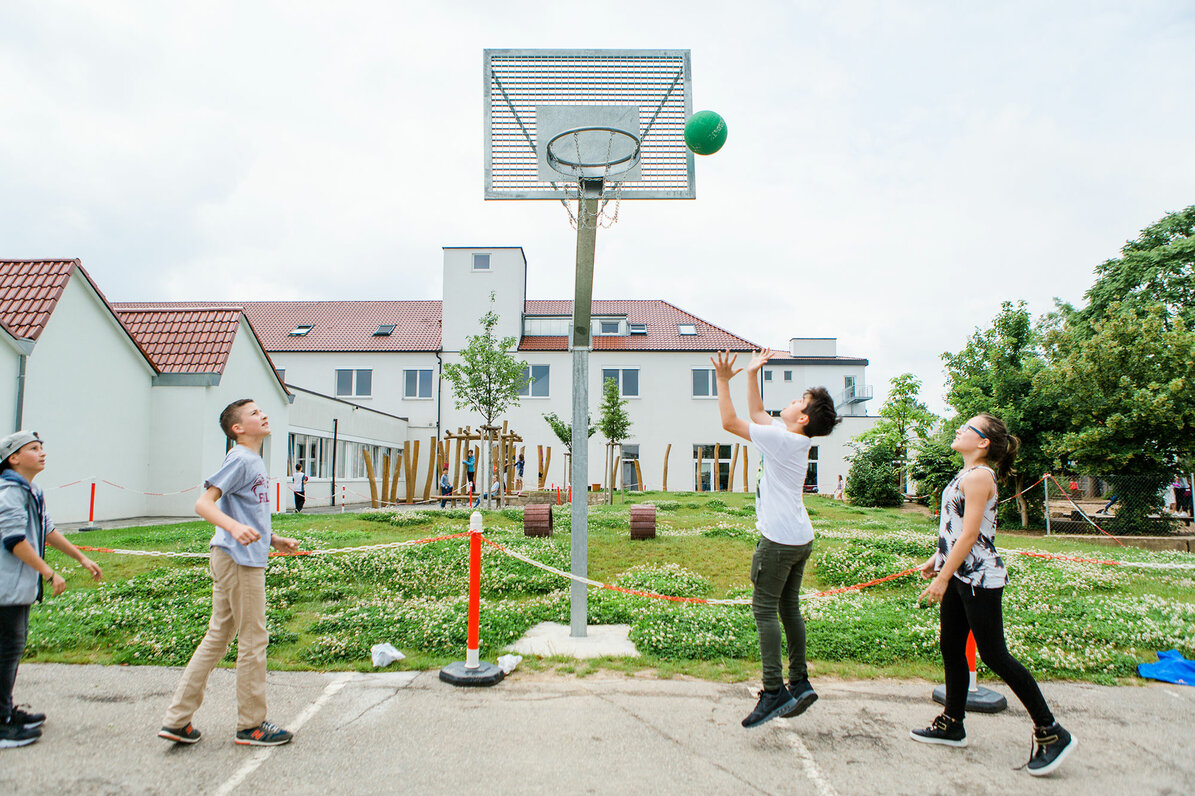 Auf dem Schulhof gibt es ein Basketballfeld, dort spielen Schüler Basketball. 