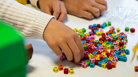 Close-up of three children's hands on a table. One hand reaches into a small pile of colourful letter beads.