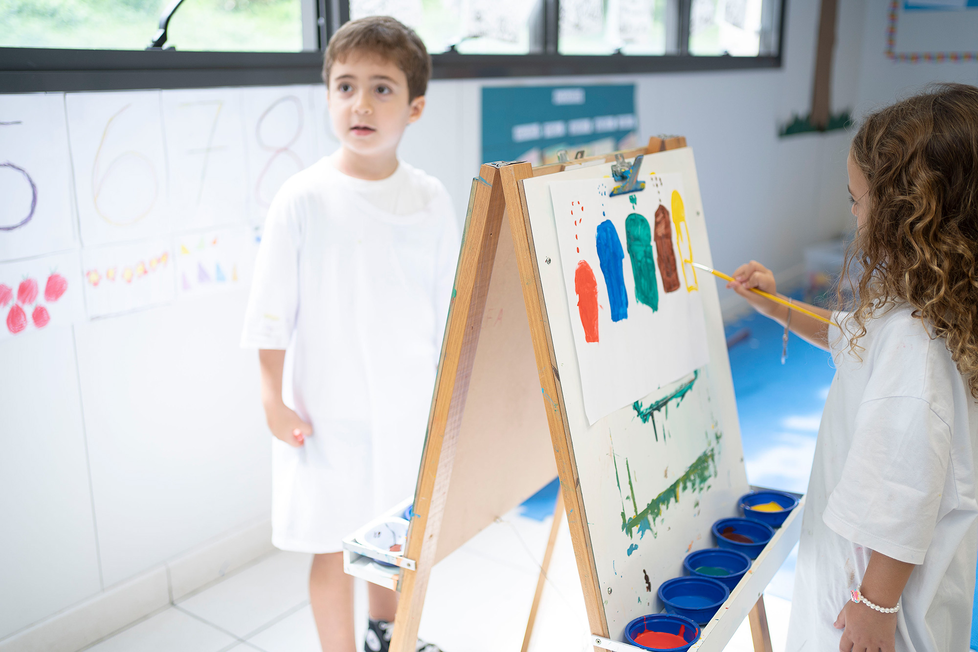 Ein Mädchen im Malkittel malt an einer Staffelei farbige Elemente mit einem Pinsel auf ein weisses Blatt Papier. 	