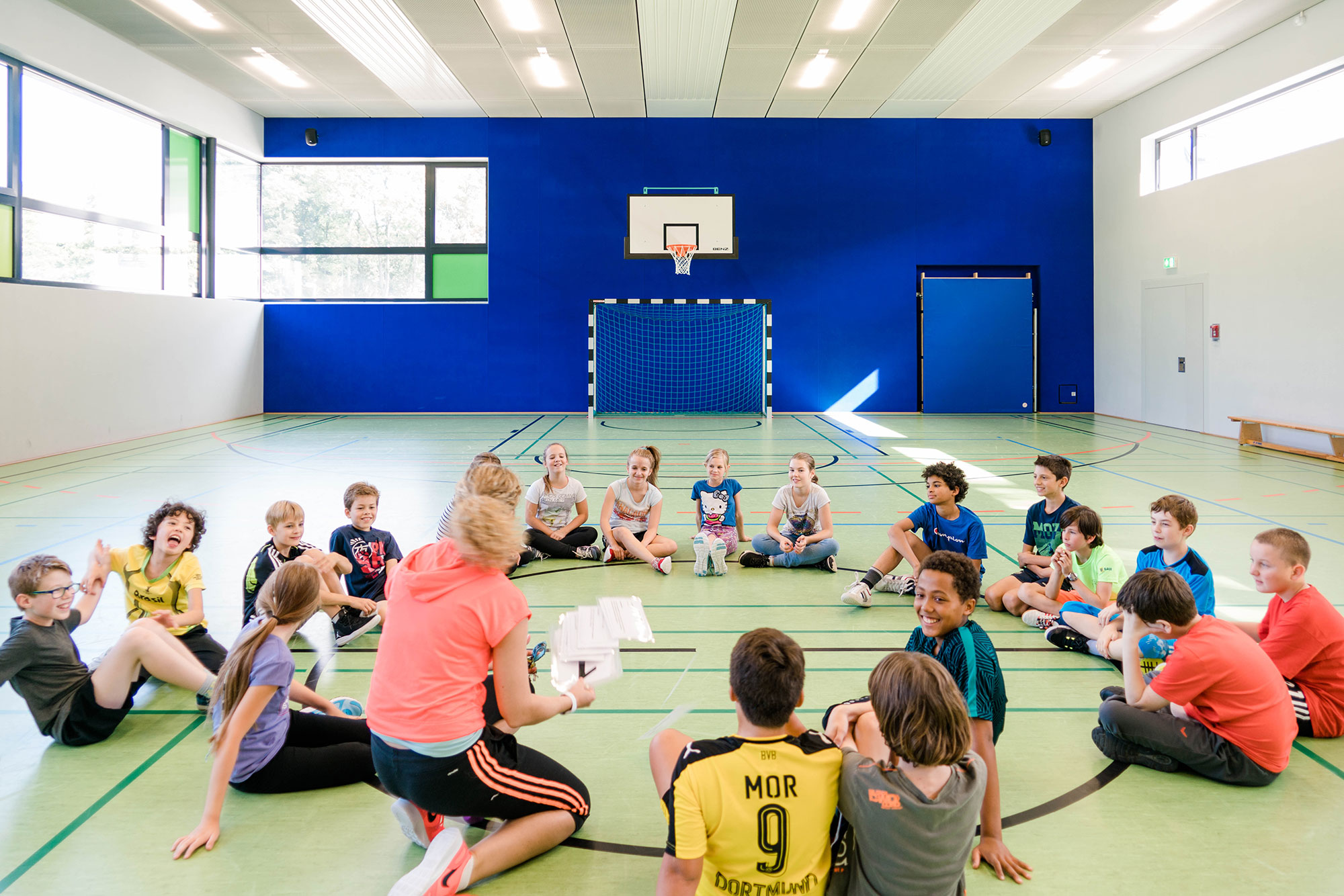 In einer modernen Turnhalle versammelt eine Sportlehrerin ihre Schülerinnen und Schüler sitzend im Kreis und gibt Instruktionen.	