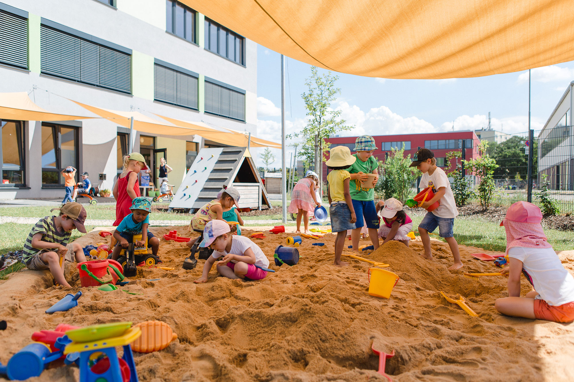 A group of kindergarten children play in the sandbox with sand toys. The sandbox is shaded by a yellow awning.