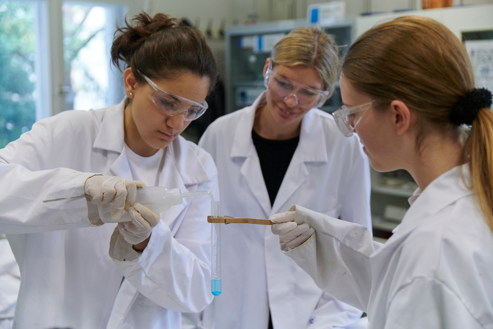 A student pours a liquid into a test tube, which another student holds with a wooden clamp. The teacher in the middle observes the process in the background.