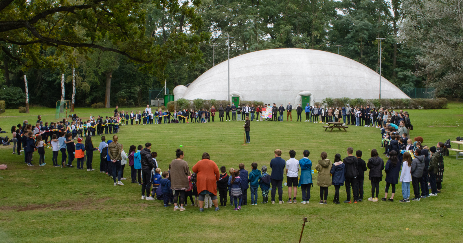 All members of SIS Berlin are standing on the lawn, forming a large circle, some of them holding a handmade paper chain. In the background you can see a white dome.