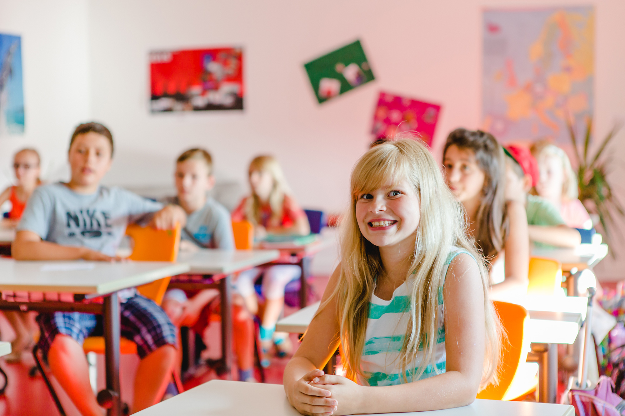 Schülerinnen und Schüler an ihren Tischen im Klassenzimmer. Im Vordergrund lächelt ein Mädchen in die Kamera.