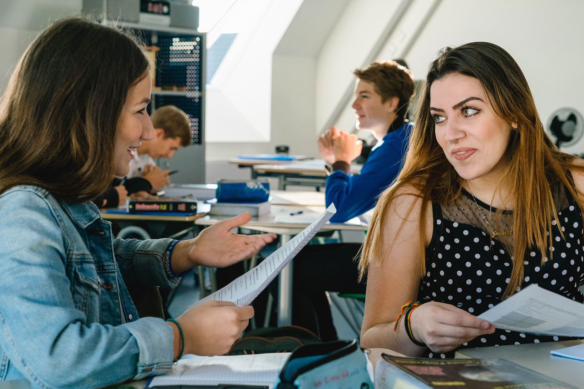 Zwei Schülerinnen sitzen am Tisch im Klassenzimmer, halten Arbeitsblätter in der Hand und sprechen. Im Hintergrund sind weitere Schüler zu sehen.