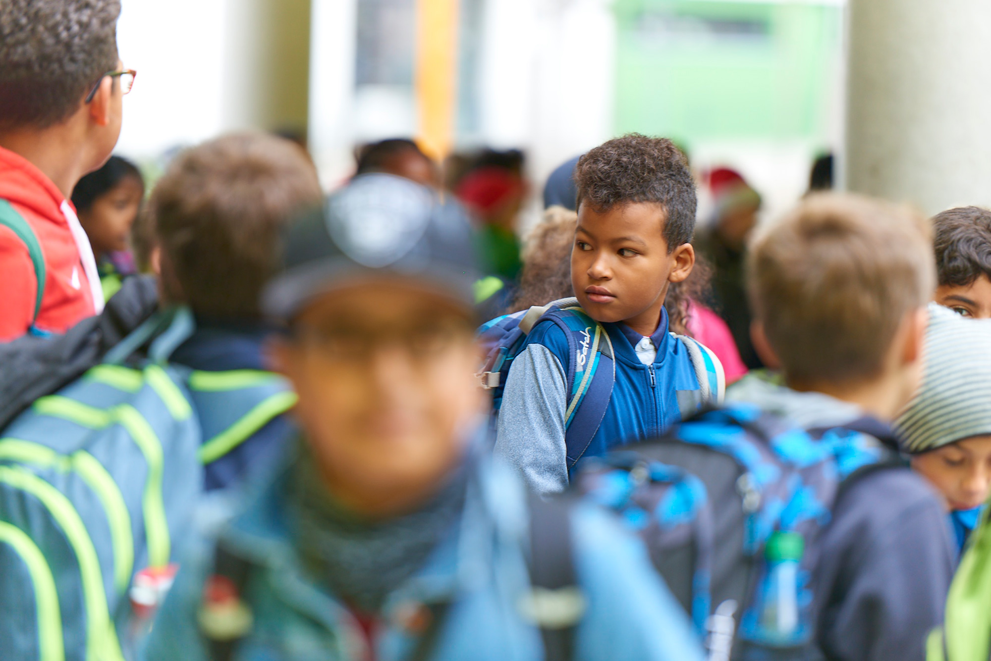 Viele Schülerinnen und Schüler mit Jacken und Schultaschen vor dem Schulgebäude. Fokus auf einem Jungen in der Mitte des Bildes.