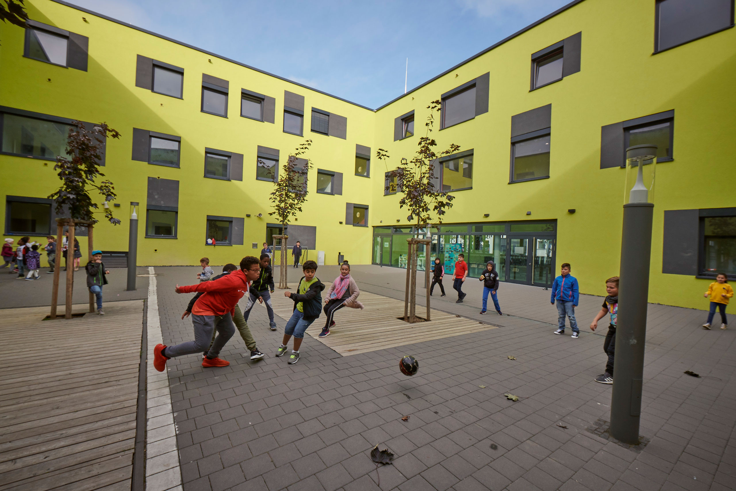 Ein Blick in den Schulhof indem Kinder am Fußball spielen sind. Rundherum ist das Schulgebäude in hellem grüngelb. 