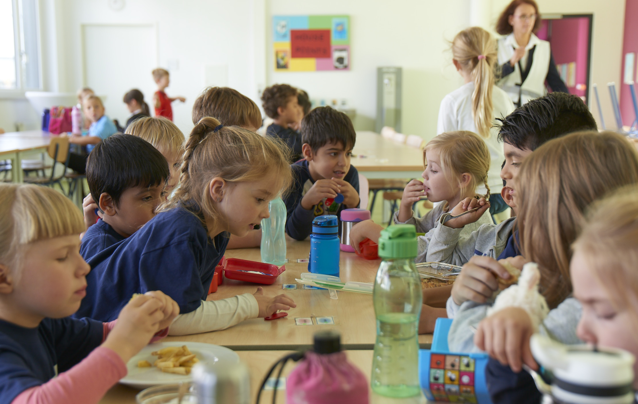 Mehrere Kinder sitzen in der Mensa an einem großen Tisch und nehmen Essen zu sich. Ein paar Kinder haben ihr eigenes Vesper dabei und die anderen essen das Essen aus der Mensa. 