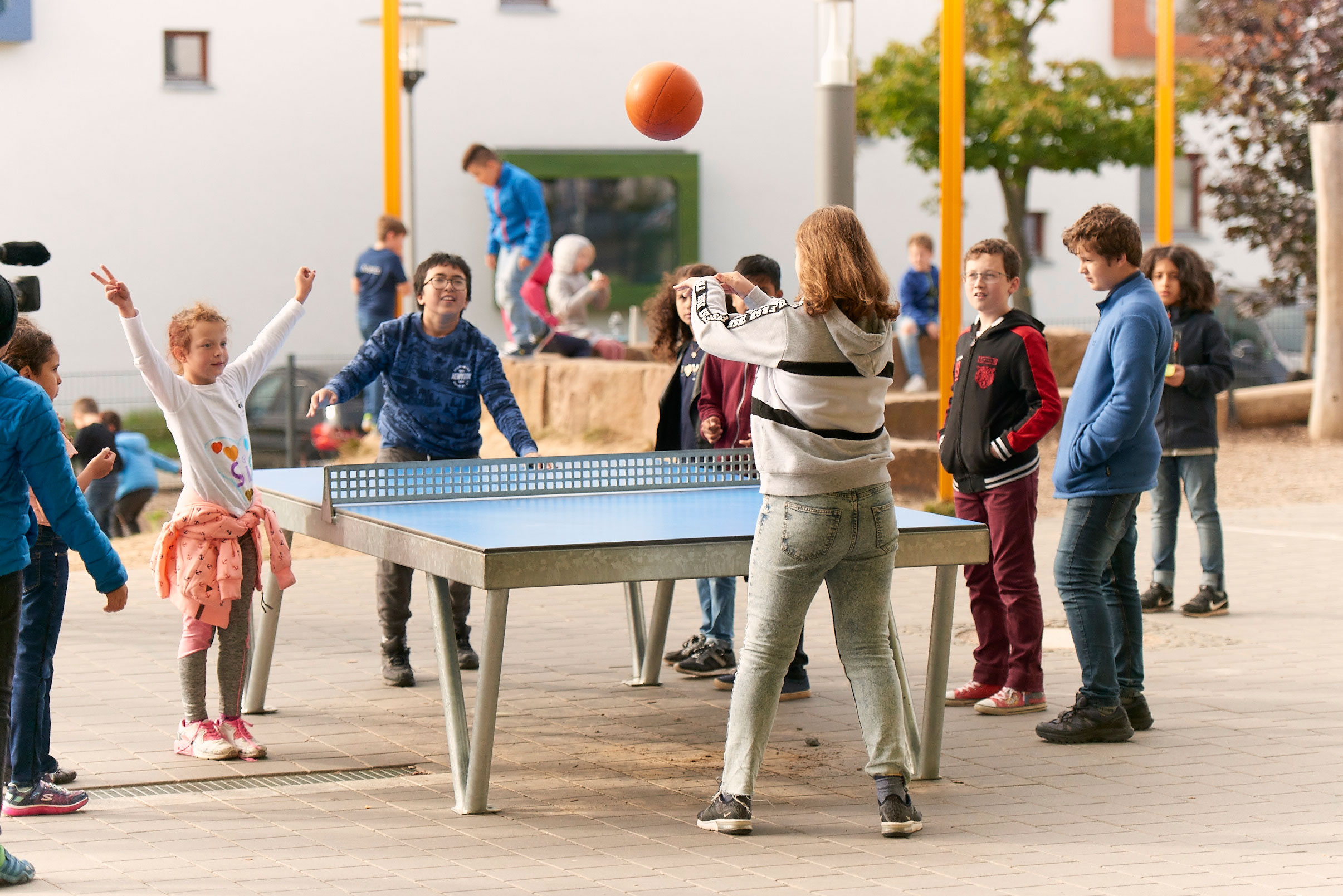 Many students are playing a game with a basketball at a table tennis table in the school yard. The ball is in the air at this moment.