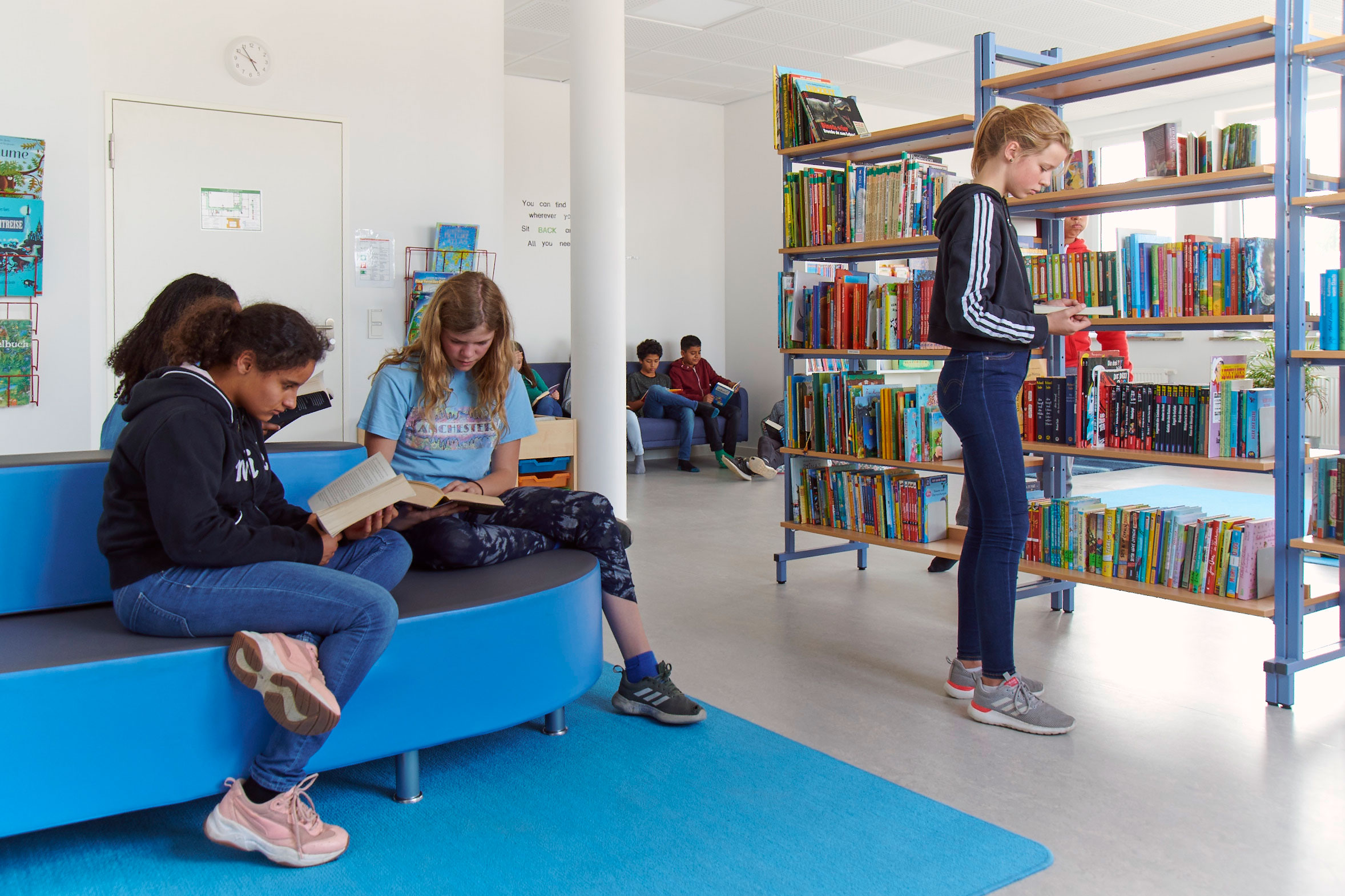 Mehrere Schülerinnen und Schüler sind in der Bibliothek und lesen in Büchern. Rechts im Bild steht ein Bücherregal.