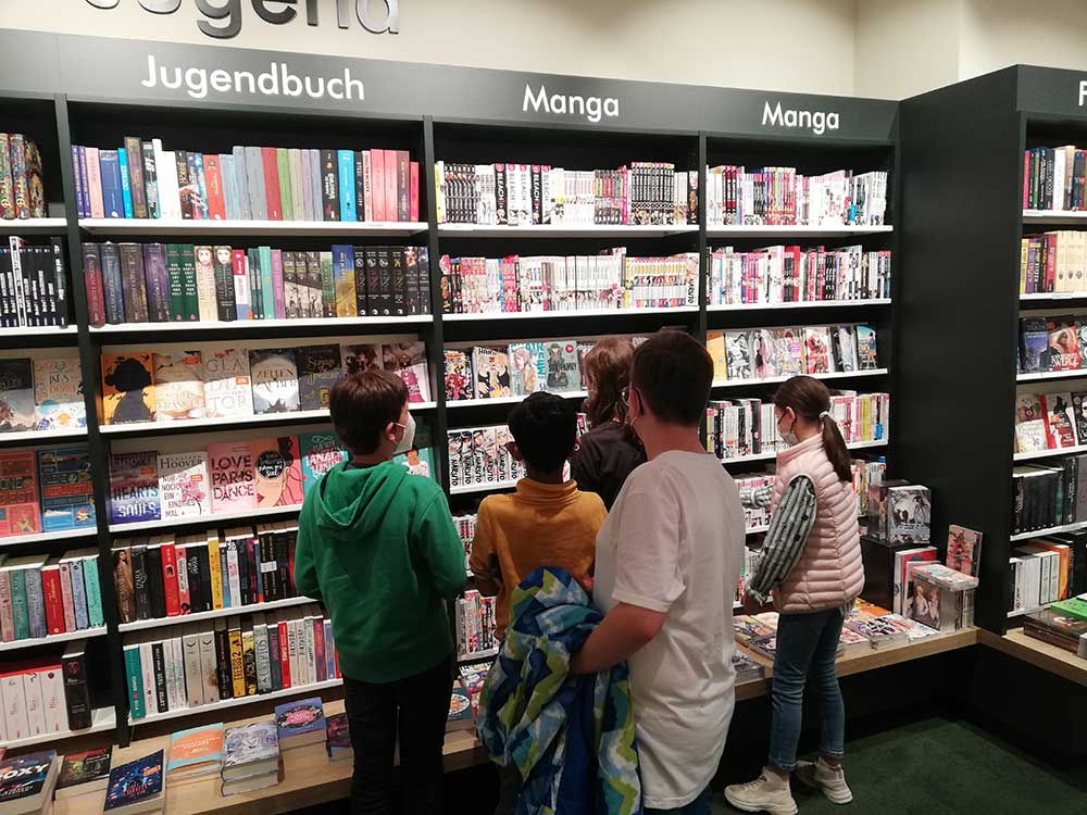 Schülerinnen und Schüler stehen vor einem Bücherregal in einem Buchladen mit dem Rücken zur Kamera.