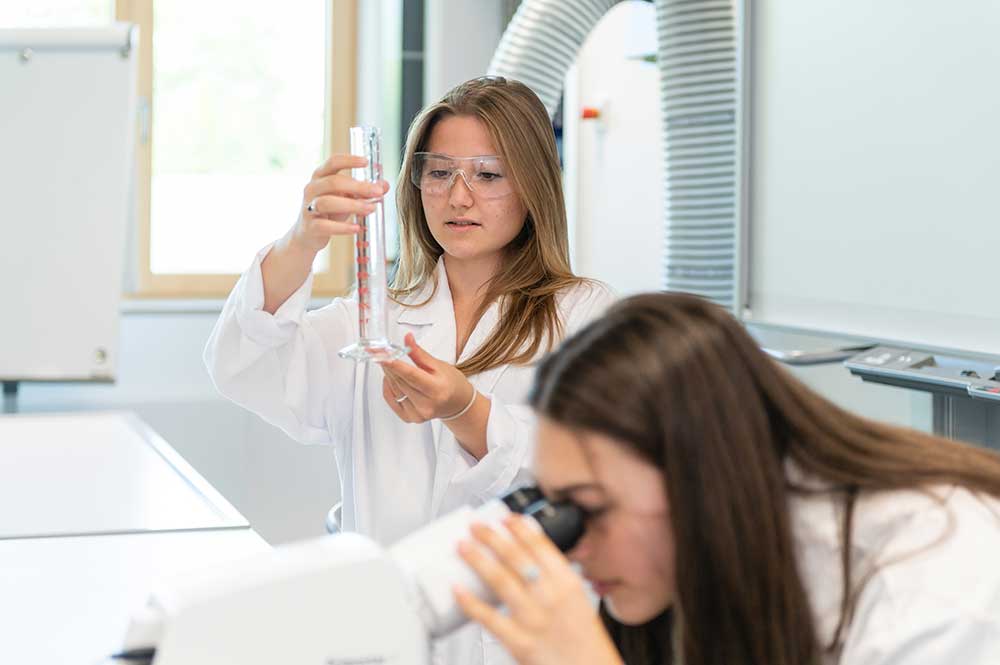 Zwei Schülerinnen sind im Labor. Eine Schülerin schaut ins Mirkoskop und die andere hält ein Reagenzglas in ihren Händen.