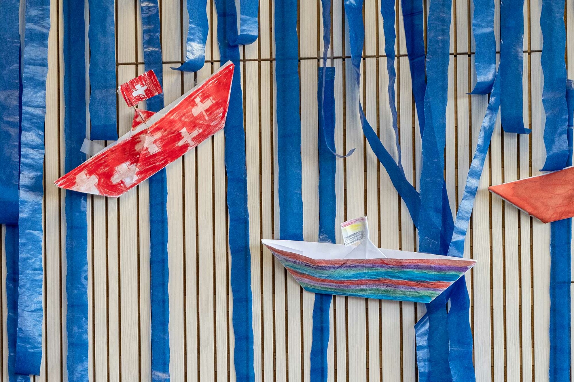 Selbst gebastelte, angemalte Papierboote hängen als Dekoration an der Wand. Eines der Boote ist mit der Schweizer Flagge bemalt.	