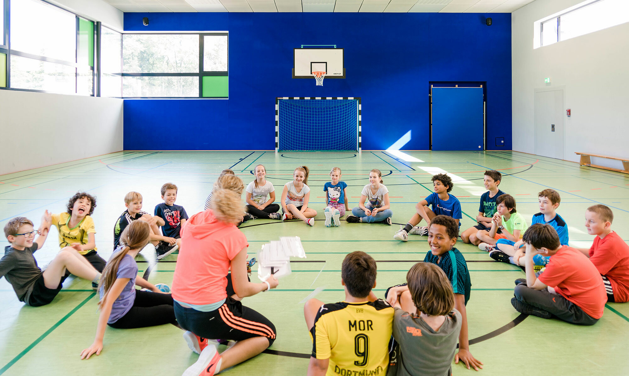 In a modern gymnasium, a sports teacher gathers her students seated in a circle and gives instructions.