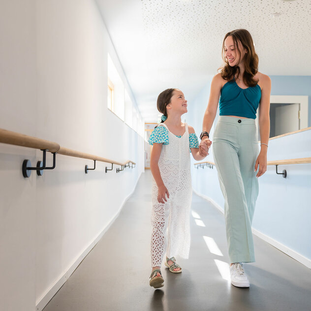 A younger and an older girl hold hands and walk through a school corridor.