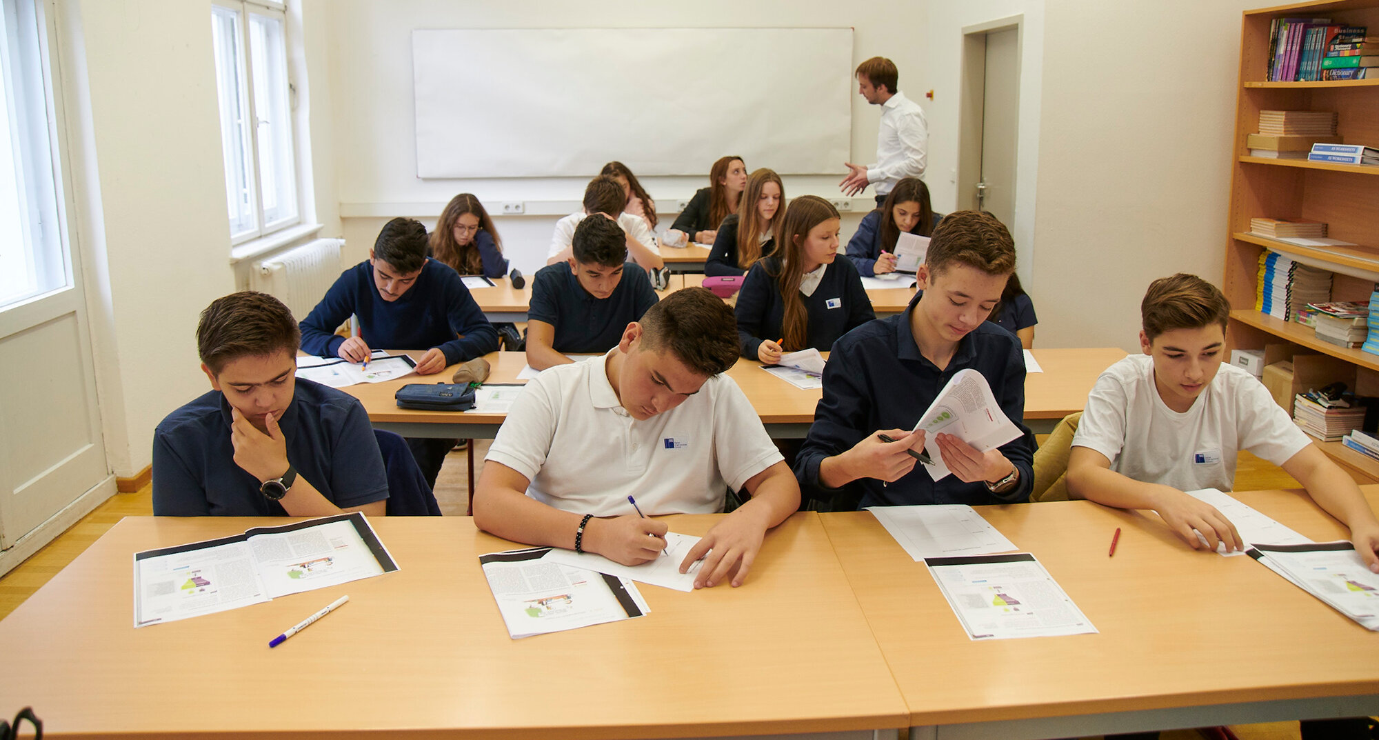 Schülerinnen und Schüler sitzen an vier Tischreihen im Klassenzimmer und bearbeiten Arbeitsblätter. 