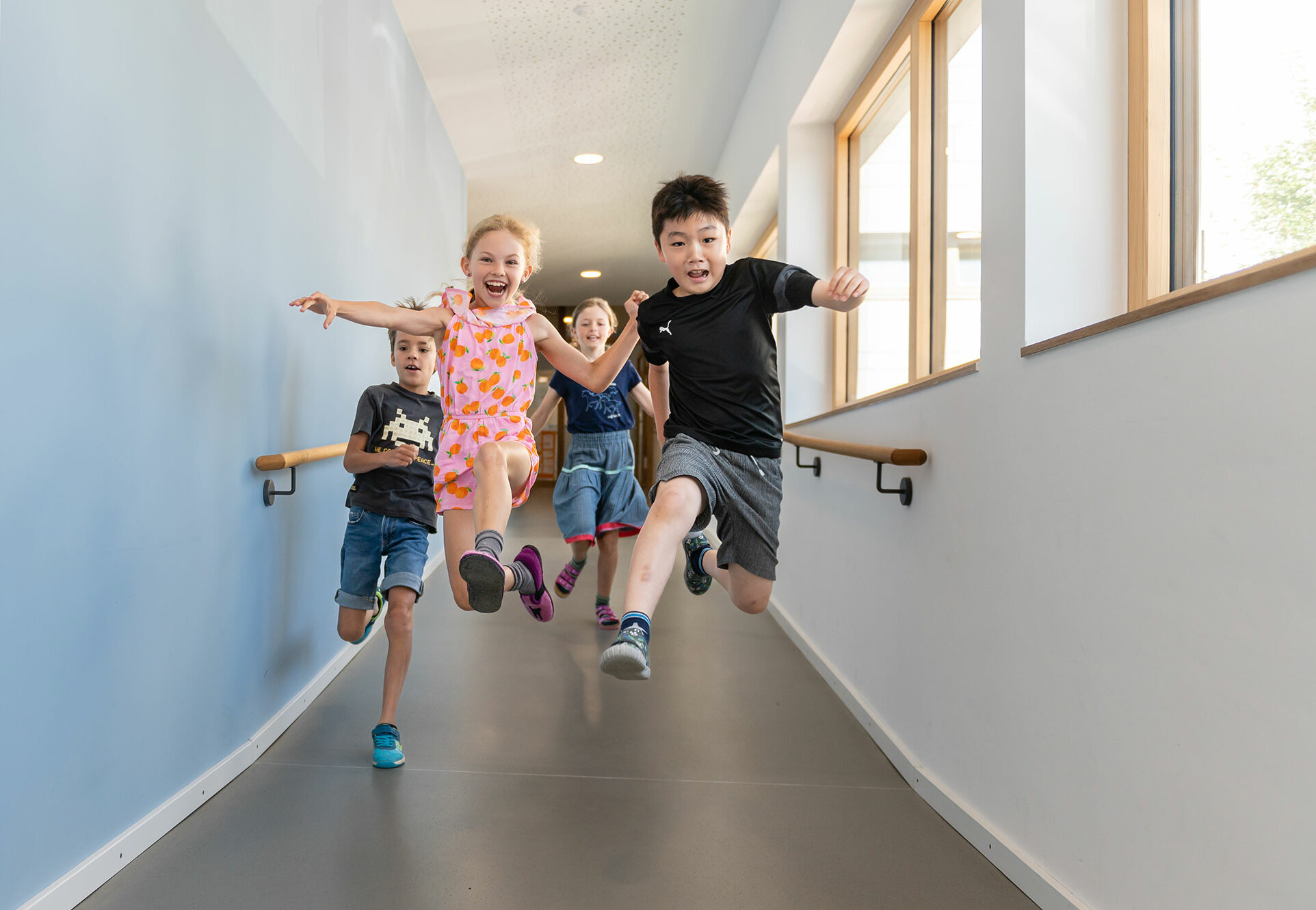 Four students can be seen running through a school corridor. The two in front make a step jump towards the camera.