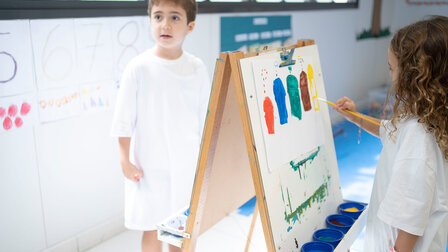 Ein Mädchen im Malkittel malt an einer Staffelei farbige Elemente mit einem Pinsel auf ein weisses Blatt Papier. 	