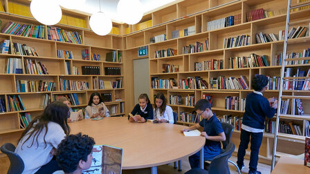 Ten children are in the library reading books at a large round table and at a small square table.