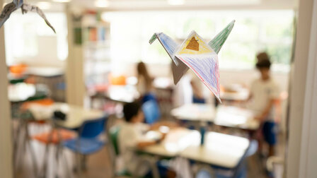Ein gebastelter, bemalter Origami-Kranich hängt im Klassenzimmer, welches unschaft in den Hintergrund tritt.	