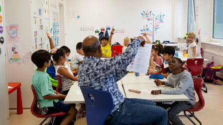 Schülerinnen und Schüler während des Unterrichts im Klassenzimmer. Ein paar halten die Arme in die Höhe.