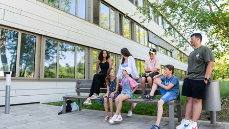 Students of different ages sit on a bench in front of the school building.