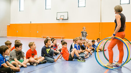 Children are sitting on the floor in the sports hall. The teacher is standing in front of them with colourful hoops in her hand.