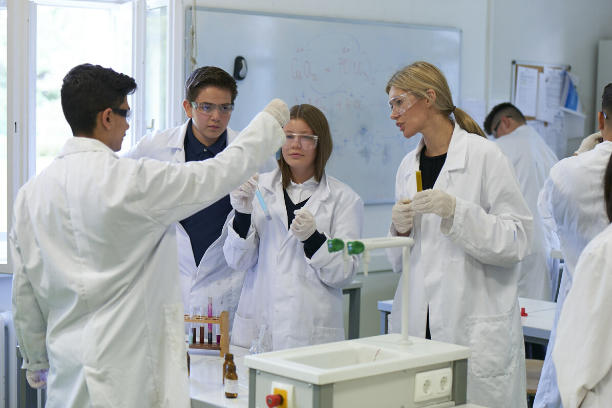 Drei Schülerinnen und Schüler und eine Lehrerin sind im Chemielabor. Sie tragen weiße Kittel und schauen auf zwei Reagenzgläser gehalten von einem Mädchen und einem Jungen.
