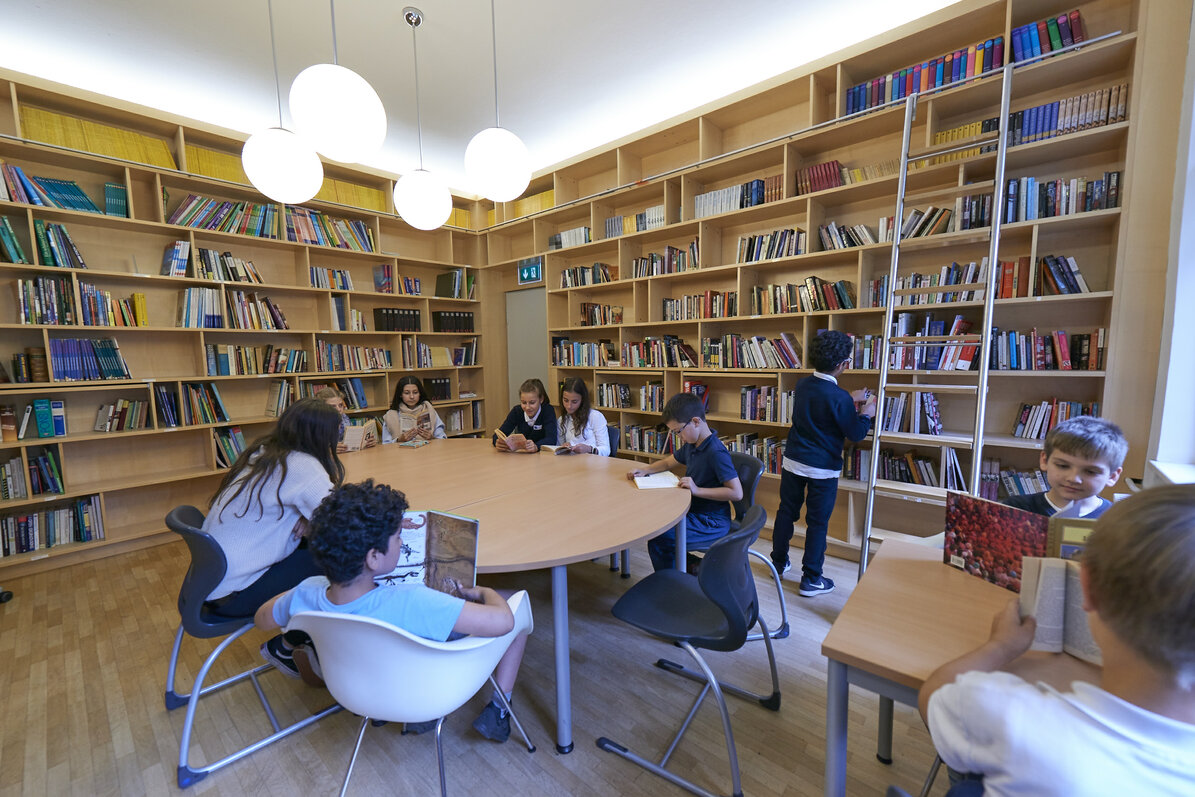 Zehn Kinder sind in der Bibliothek und lesen Bücher an einem großen runden Tisch und an einem kleinen eckigen Tisch.