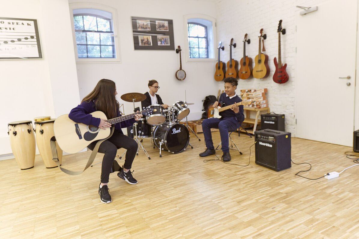 Three students make music together. One student plays acoustic guitar, one student plays drums, one students plays electric guitar. There are more acoustic guitars hanging on the wall.