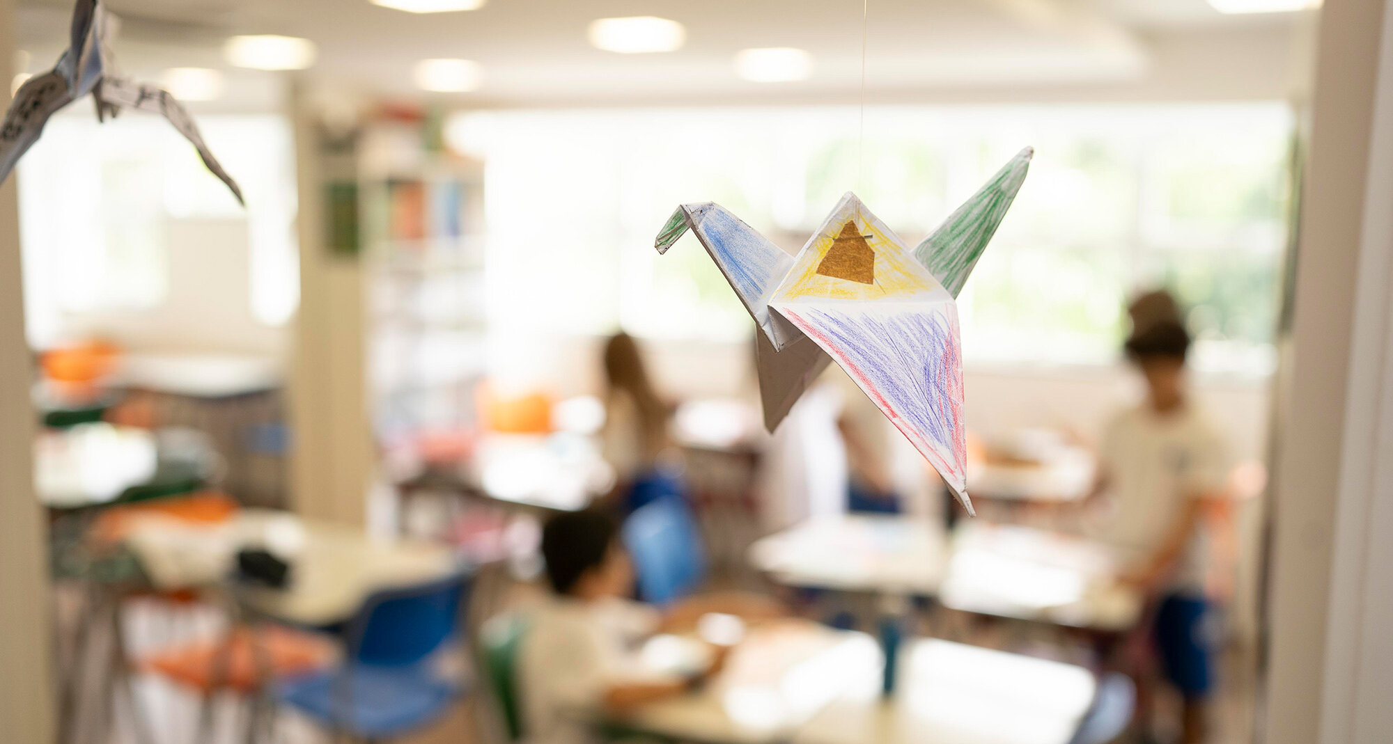 Ein gebastelter, bemalter Origami-Kranich hängt im Klassenzimmer, welches unschaft in den Hintergrund tritt.	