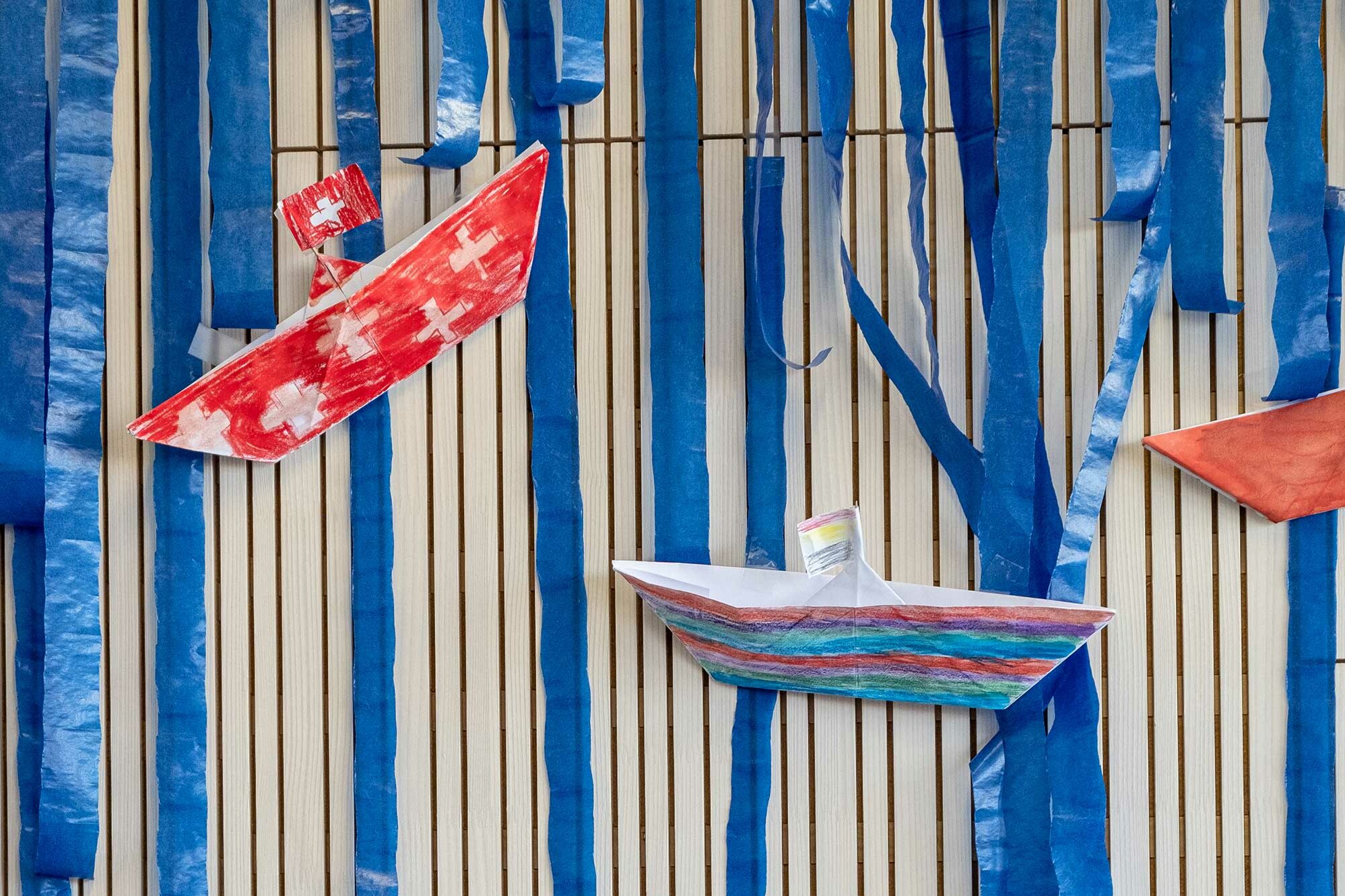 Self-made, painted paper boats hang on the wall as decoration. One of the boats is painted with the Swiss flag.	