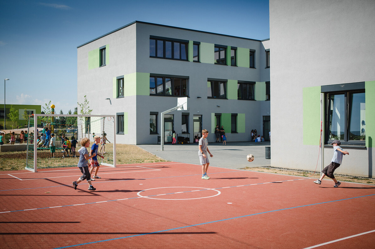 Vor dem Gebäude ist ein kleiner Sportplatz auf dem mehrere Schüler Fußball spielen.