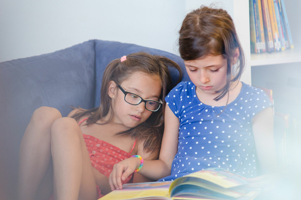 Two girls are sitting on a sofa reading a book together. One girl leans against the other.
