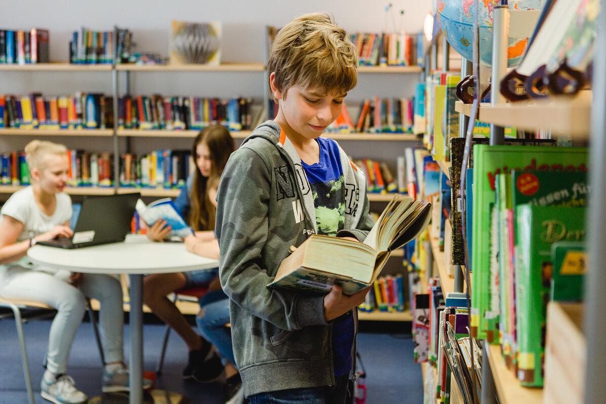 Ein Schüler ist in der Bibliothek und steht mit einem offenen Buch in der Hand vor einem gefüllten Bücherregal. Hinter ihm ist ein weiteres volles Bücherregal und ein Tisch an dem ein paar Schülerinnen mit ihren Laptops sitzen. 