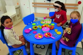 Drei Mädchen sitzen am Kindertisch mit Spielzeug-Geschirr, -Obst und -Gemüse