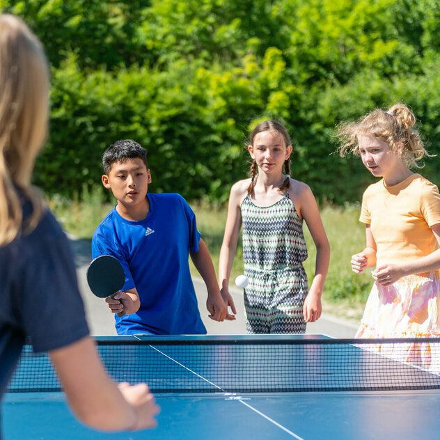 Four children are standing around a table tennis table. Two of them are playing table tennis.