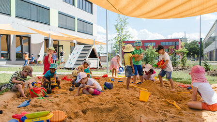 A group of kindergarten children play in the sandbox with sand toys. The sandbox is shaded by a yellow awning.