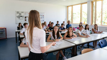 Schülerinnen und Schüler sitzen an ihren Plätzen im Klassenzimmer und schauen nach vorne zur Lehrerin.