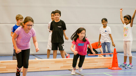 A group of primary school children are in the sports hall. A folded long bench marks a waiting area. Two girls in the foreground are just starting a race.