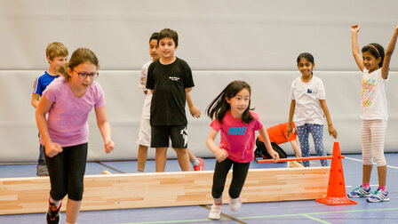 A group of primary school children are in the sports hall. A folded long bench marks a waiting area. Two girls in the foreground are just starting a race.