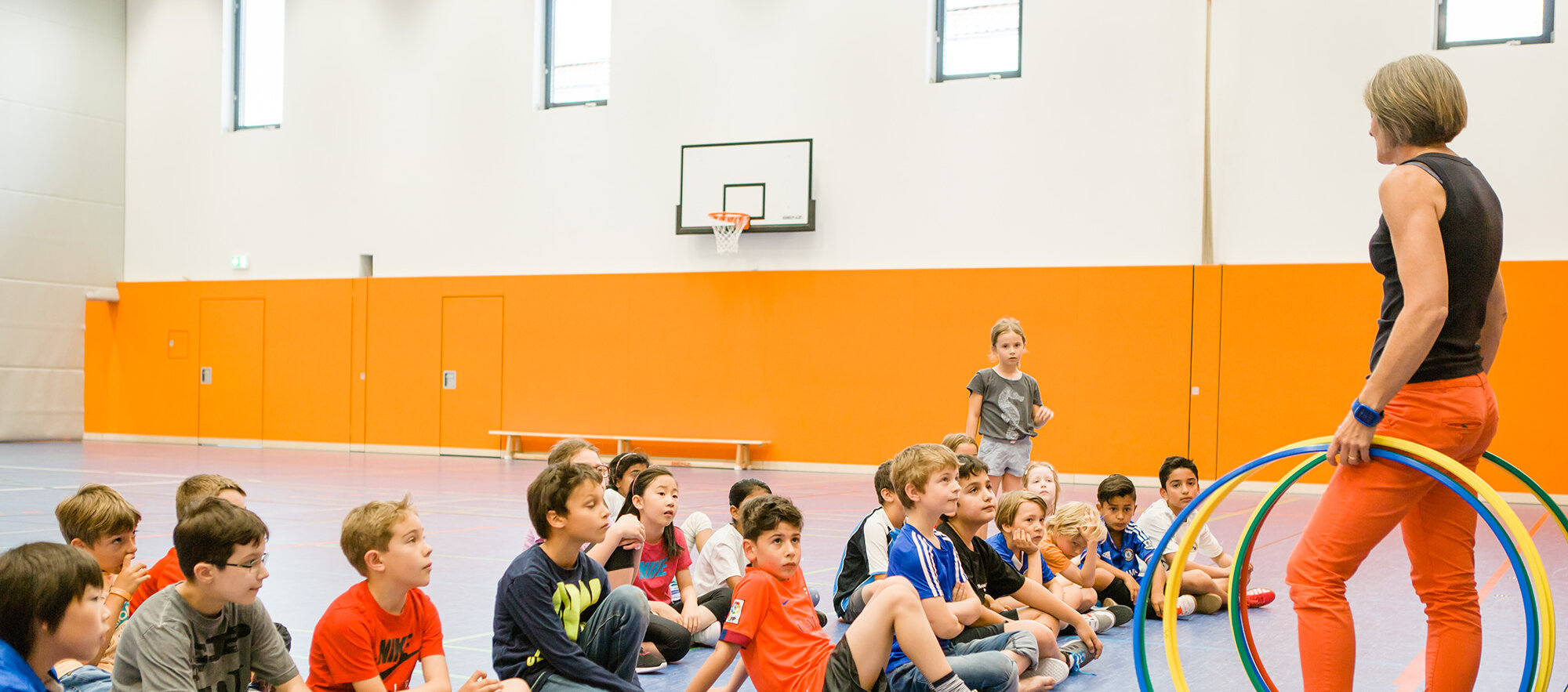 Children are sitting on the floor in the sports hall. The teacher is standing in front of them with colourful hoops in her hand.