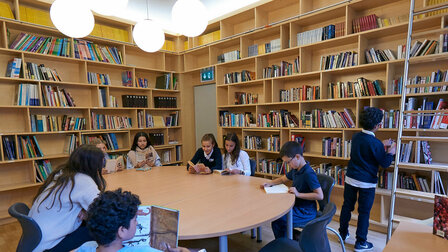 Ten children are in the library reading books at a large round table and at a small square table.