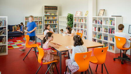 View of the school library. Children are reading, picking up books or sitting at the computer.