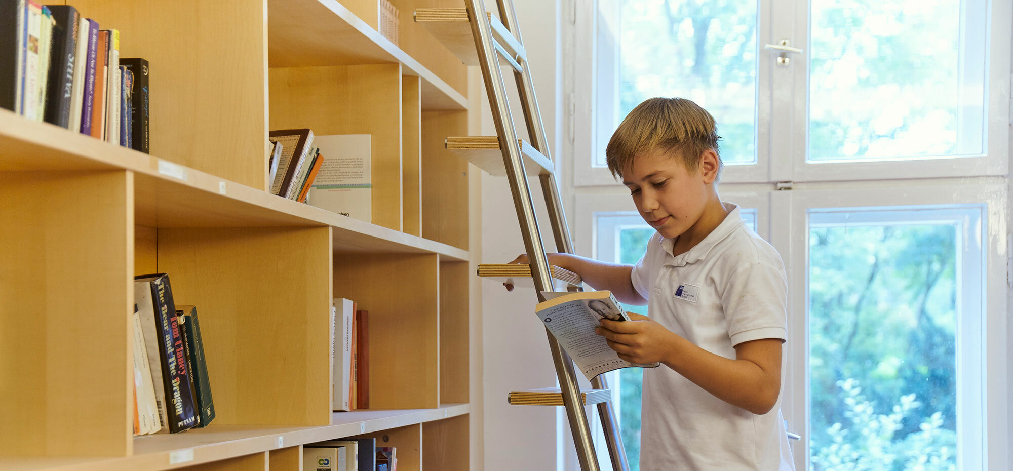 Ein Junge steht in der Schulbibliothek auf einer Leiter und holt sich ein Buch.	